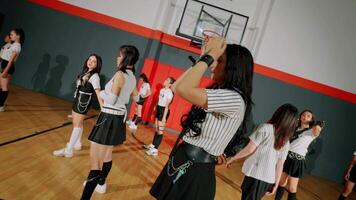 A group of dancers in a gymnasium, wearing stylish outfits with stripes and skirts. They are posing confidently, showcasing their dance moves. The background features a basketball hoop video