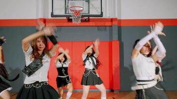 A young woman performing a dance routine in a basketball court setting. She wears a striped shirt and a black skirt with chains, showcasing energetic movement. The background features basketball video