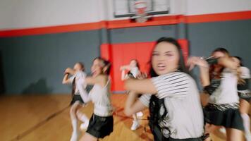 A group of four dancers performing in a gymnasium, wearing matching striped outfits with skirts. The lead dancer is in focus, showcasing energetic dance moves, while the others follow suit video