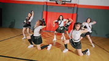 A dynamic dance performance in a gymnasium featuring multiple dancers in stylish outfits. The background includes a basketball hoop and colorful wall panels, creating an energetic atmosphere. video