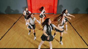 A group of six young women performing a dance routine in a gymnasium. They are dressed in matching outfits with skirts and sneakers, showcasing energetic movements. The background features a red wall video