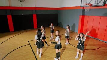 A group of six girls in matching outfits practicing a dance routine in a basketball gym. The scene captures their movements from behind, showcasing the gym's wooden video