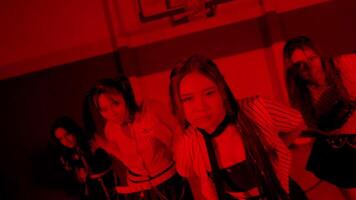 A group of four young women dressed in stylish outfits, posing dramatically in a dimly lit gymnasium with red lighting. The atmosphere is intense and energetic, showcasing a dance or performance vibe. video