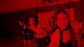 A dynamic dance performance in a gymnasium, featuring dancers in striped outfits. The scene is illuminated with red lighting, creating a dramatic atmosphere. The dancers are captured in motion video