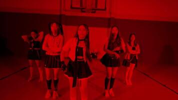 A group of six young women performing a dance routine in a gymnasium. The lighting is predominantly red, creating a dramatic atmosphere. They wear stylish outfits, showcasing a mix of sporty video