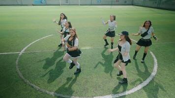 A group of six young women performing a dance routine on a soccer field. They are dressed in sporty outfits, showcasing energetic movements. The green turf and goalposts are visible in the background. video
