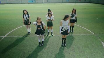 A group of six young women performing a dance routine on a sports field. They are dressed in stylish outfits, showcasing a mix of skirts and tops, with a vibrant green turf background. video