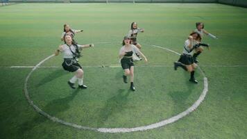 A group of six dancers performing a synchronized routine on a soccer field. They are dressed in sporty outfits, showcasing energetic poses with arms raised. The green turf and circular field markings video