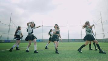 A group of six dancers performing on a grassy field, dressed in stylish black outfits. The scene is set against a cloudy sky, with a sports net in the background. video