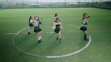 A group of six dancers performing in a choreographed routine on a green sports field. They are dressed in stylish outfits, showcasing a mix of black and white colors, with some wearing gloves video