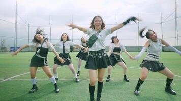 A group of five dancers performing a choreographed routine on a green sports field. They are dressed in stylish outfits, showcasing synchronized movements against a cloudy sky. video