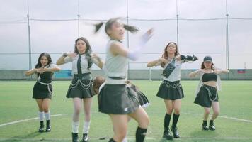 A group of young women performing a dance routine on a sports field. They are dressed in stylish outfits, with one woman in the foreground energetically pointing at the camera video