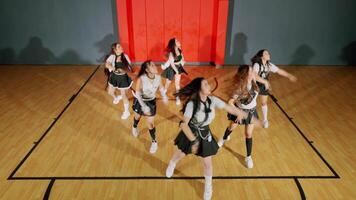 A group of six dancers performing a choreographed routine in a gymnasium. They are dressed in stylish outfits, showcasing energetic movements against a backdrop of a red wall and wooden floor. video