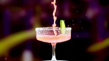 A pink margarita is poured into a glass with a lime garnish, set against a colorful, blurred background video