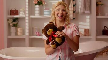 Smiling joyful Caucasian girl female teenager woman in pink candy glamour bathroom looking at camera holding Teddy Bear playing showing fluffy toy happy carefree indoors present gift valentine's day photo