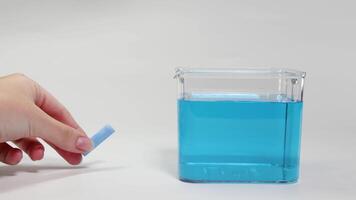 A hand drops chalk into a water-filled container, observing its sinking behavior under controlled lighting video