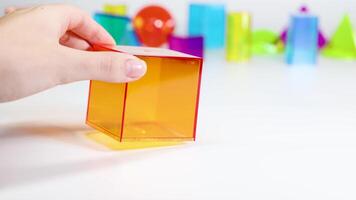 A hand manipulates a transparent orange cube among various colorful geometric shapes on a white surface video