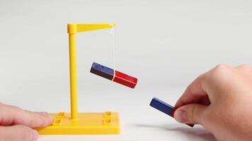 Hands manipulate magnets to demonstrate attraction and repulsion using a suspended bar magnet on a yellow stand video