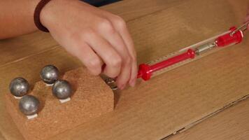 Hands manipulate a spring scale and weights on cardboard to demonstrate friction and force measurement in a controlled setup video