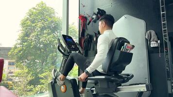 A man uses a recumbent bike in a well-lit gym with large windows and a view of trees video