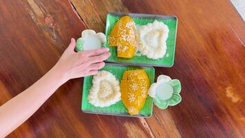 A hand pours coconut milk over mango sticky rice on a wooden table in a bright, tropical setting video