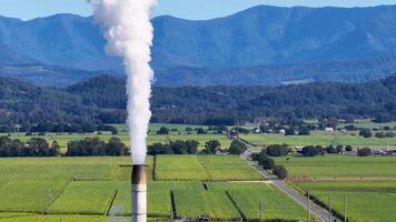 Aerial footage captures steam rising from a sugarcane factory amidst lush fields and distant mountains under clear blue skies video