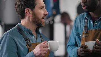 Multiethnic coffee factory two workers drinking beverage tasting discussing flavor aroma strength blend warmth work break Caucasian African men talk drink espresso barista resting roast manufacturing video