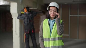 Happy multinational builders Asian woman engineer mobile phone call communication smartphone construction coordination safety helmet planning Caucasian man tools drilling noise distraction renovation video