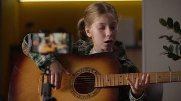 Little girl blogger recording mobile phone blog playing guitar online lesson Caucasian teenager child kid talented student learning play music song teaching streaming smartphone camera tripod at home video