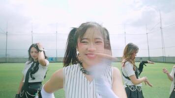 A group of young women performing a dance routine on a sports field. The foreground features a smiling girl with pigtails, wearing a striped outfit video