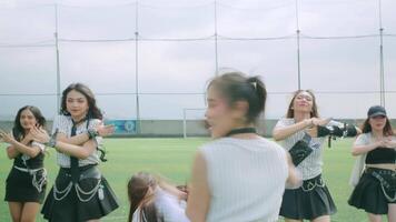 A group of dancers performing on a sports field, showcasing dynamic movements. The foreground features a dancer with a playful expression, while others are in motion behind her video