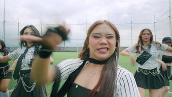 A dynamic dance performance by a group of five young women on a green turf field. They are dressed in stylish outfits, showcasing energetic movements against a cloudy sky backdrop. video