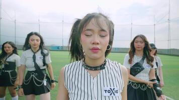 A group of young women performing a dance routine on a sports field. The central figure has pigtails and wears a striped shirt with a choker, while others are in matching outfits video
