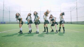 A group of six female dancers performing a choreographed routine on a green turf field. They are dressed in stylish outfits with a mix of black and white video
