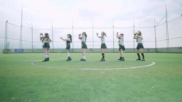 A group of six girls performing a synchronized dance on a sports field. They are dressed in matching outfits, showcasing their choreography against a backdrop of a cloudy sky. video