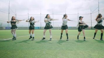 A group of six dancers performing a choreographed routine on a green turf field, surrounded by a netted enclosure. They are dressed in stylish black and white outfits video