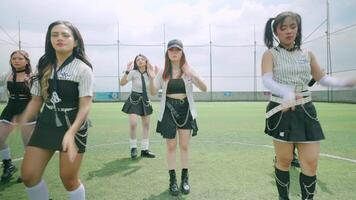 A group of five female dancers performing on a soccer field, wearing stylish black and white outfits with chains and accessories. They are in a dynamic dance pose under a bright sky. video