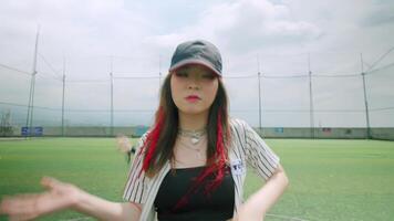 A lively scene on a sports field featuring a young woman dancing energetically. She wears a cap and a striped shirt, with a playful expression. In the background, other dancers are visible video