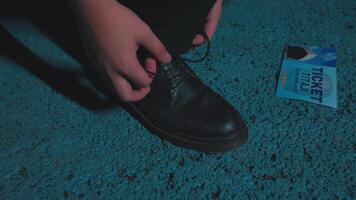 A close-up of a person's hands tying shoelaces on a black shoe, with a blue ticket lying on a textured surface nearby. video