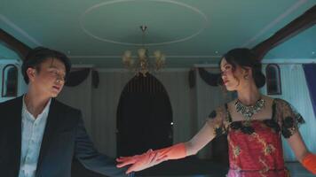 A couple dancing in an elegant, dimly lit ballroom. The woman wears a red and black dress with intricate patterns, while the man is in a formal suit video