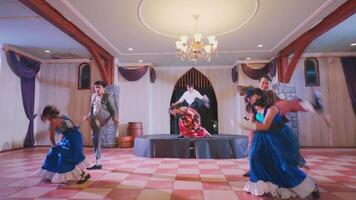 A lively dance performance featuring six dancers in colorful costumes on a stage. The setting includes elegant decor with a chandelier and wooden accents, creating a festive atmosphere. video
