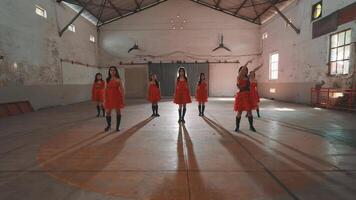A group of dancers in red dresses performing in an empty industrial space. The dancers are in various poses, showcasing their choreography. The setting features high ceilings and large windows video