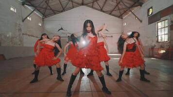 A group of young dancers performing in a spacious warehouse. They are wearing vibrant red dresses and black boots, showcasing energetic dance moves. The industrial setting adds a unique backdrop video