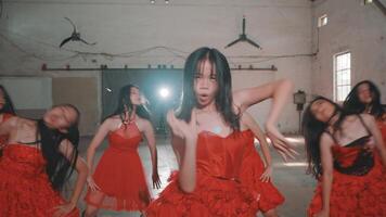 A group of four dancers in red dresses performing in an industrial space. The dancers showcase energetic movements, with a focus on rhythm and style. The background features large fans video