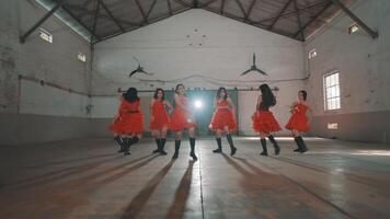A group of five dancers in red dresses performing in an industrial space. The dancers are in a dynamic pose, showcasing their choreography. The background features a large open area video