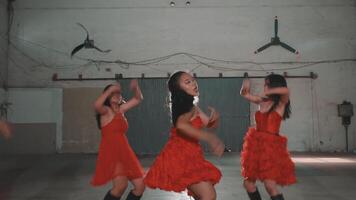 A group of young women in red dresses performing a dance routine in an industrial space. The atmosphere is energetic and vibrant, with bright lighting highlighting their movements. video