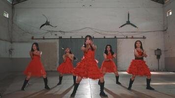 A group of young women in red dresses performing a dance routine in an industrial setting. The dancers are showcasing energetic movements, with a focus on rhythm and coordination. video