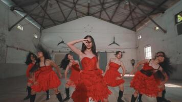 A group of six dancers in red dresses performing in an industrial space. The dancers showcase synchronized movements, with a focus on energy and style. The setting features high ceilings video