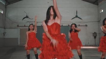 Three dancers in red dresses performing energetically in an industrial space. The focus is on the central dancer, who is expressing intense emotion. The background features a simple, rustic setting video