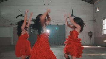 A group of young women wearing vibrant red dresses, dancing energetically in a spacious indoor setting. Their expressions show joy and excitement as they perform together. video